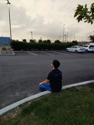 Matthew, age 21, sits on the curb of a hotel parking lot in Indianapolis. He is cross-legged with his camera strap slung over his left shoulder, as he peers intently into a gray sky, ready for an approaching thunderstorm..